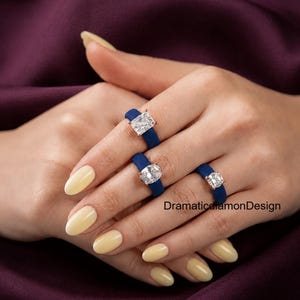 May include: Three rings featuring dark blue bands and clear, faceted stones in rectangular, oval, and round shapes, set in metal prongs. The text DramaticDiamonDesign is visible on the right side of the image, near the bottom ring.