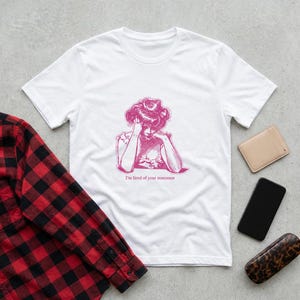 May include: White t-shirt with a pink illustration of a woman resting her head on her hands. The text "I'm tired of your nonsense" is below the illustration. A red and black plaid shirt, a beige wallet, a black phone, and a leopard print glasses case are also visible.