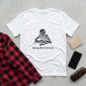 May include: White t-shirt featuring a black graphic of a skeleton in a meditative pose, with the text "Waiting Since Eternity" below. A red and black plaid shirt is partially visible on the left. Other items include a phone, wallet, and glasses case.