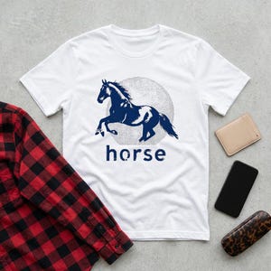 May include: White t-shirt featuring a navy blue horse graphic inside a gray circle, with the word "horse" below. A red and black plaid shirt, a beige wallet, a black phone, and a leopard print glasses case are also visible.