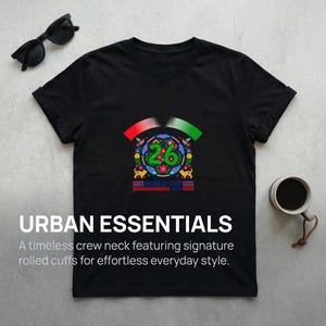 May include: Black crew neck t-shirt with a colorful graphic featuring the number 26 and the words "WORLD CUP". The shirt also has the text "URBAN ESSENTIALS" and the description "A timeless crew neck featuring signature rolled cuffs for effortless everyday style."