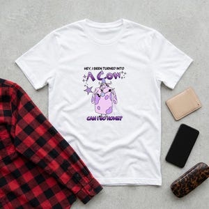 May include: White t-shirt with a purple and pink cow graphic and the text "HEY, I BEEN TURNED INTO A COW CAN I GO HOME?". The shirt has a crew neck and short sleeves. A red and black plaid shirt is partially visible.