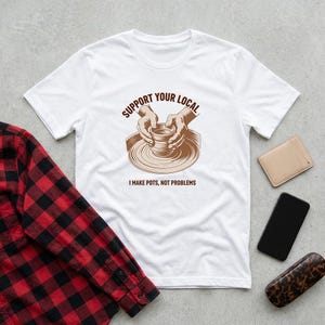 May include: White t-shirt with a brown graphic of hands shaping a pot on a pottery wheel. The text "SUPPORT YOUR LOCAL" arches above the image, with "I MAKE POTS, NOT PROBLEMS" below. A red and black plaid shirt, a tan wallet, a black phone, and a leopard print glasses case are also visible.
