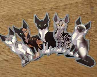 May include: Five cat stickers with various designs and colors. The stickers feature cats in shades of black, brown, gray, and white, with detailed patterns and blue or green eyes. The stickers are arranged on a wooden surface.