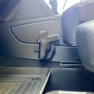 May include: Black leather holster mounted inside a vehicle, holding a silver and black handgun. The holster is attached to the vehicle's interior panel near the seat. A black floor mat is visible in the foreground.