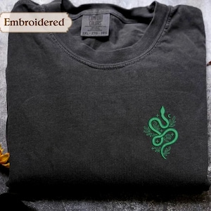 May include: A folded dark grey t-shirt with a green embroidered snake design. The shirt has a crew neck and short sleeves. The tag reads "Comfort Colors". The word "Embroidered" is on a banner in the upper left corner.