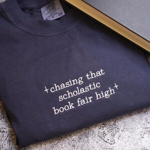 May include: A navy blue sweatshirt with the text "+ chasing that scholastic book fair high +" in white. A closed book with a gold edge is next to the sweatshirt. The sweatshirt is folded on a textured surface.