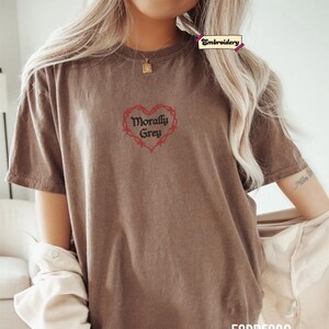 A woman wearing a brown t-shirt with a heart-shaped embroidered design and the text "Morally grey" printed on it.