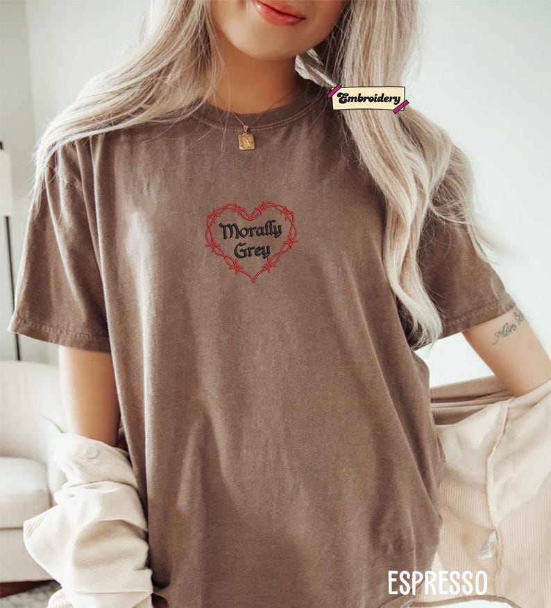 A woman wearing a brown t-shirt with a heart-shaped embroidered design and the text "Morally grey" printed on it.