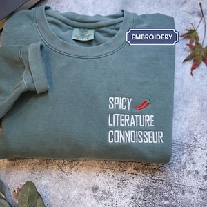 May include: A sage green sweatshirt with rolled sleeves. The front displays the embroidered text "SPICY LITERATURE CONNOISSEUR" and a red chilli pepper graphic. The word "EMBROIDERY" is in a blue box.