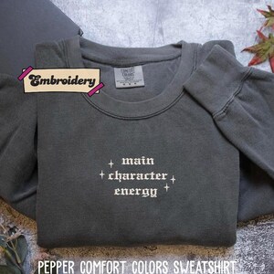 A gray sweatshirt with the text "main character energy" embroidered on it, along with a label that says "Embroidery" and a small leaf decoration.