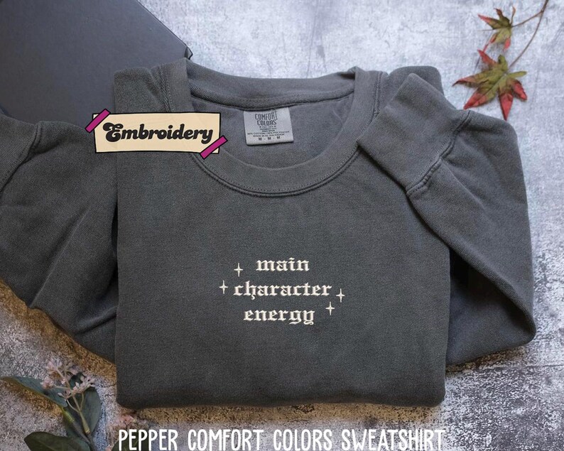 A gray sweatshirt with the text "main character energy" embroidered on it, along with a label that says "Embroidery" and a small leaf decoration.