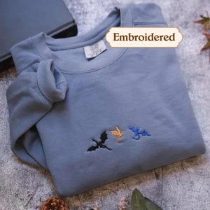Peut inclure: Un sweat-shirt à col rond bleu ardoise orné de trois petites silhouettes de dragons brodées en noir, orange et bleu. Une étiquette rectangulaire portant le texte Embroidered est placée au-dessus du centre de la poitrine du vêtement.