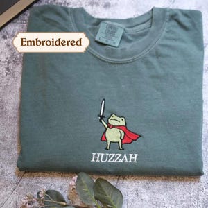 a green t-shirt with a cartoon frog wearing a red cape and the word "HUZZAH" printed on it. The t-shirt is embroidered with a small frog character, and there is a leaf and a tag visible in the background.