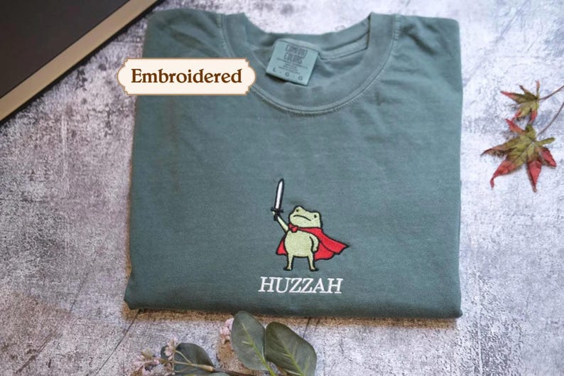 a green t-shirt with a cartoon frog wearing a red cape and the word "HUZZAH" printed on it. The t-shirt is embroidered with a small frog character, and there is a leaf and a tag visible in the background.