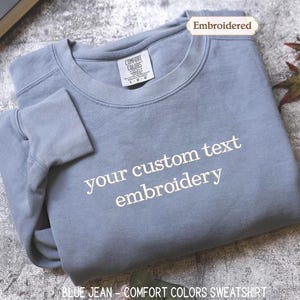 May include: A blue jean Comfort Colors sweatshirt with the text "your custom text embroidery" embroidered in white. The sweatshirt is folded and has a tag that reads "Comfort Colors".