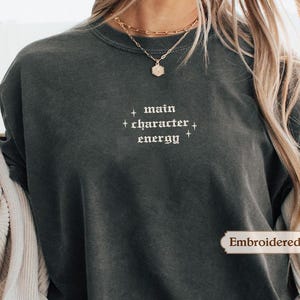A person wearing a gray sweatshirt with the text "main character energy" printed on it.