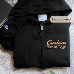 May include: Black zip-up hoodie with a hood and drawstring. The hoodie has the Champion logo on the sleeve and the text "Custom Text or Logo" embroidered in gold. The word "Embroidered" is also displayed.