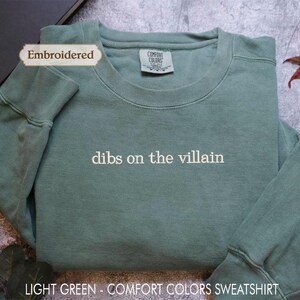 A light green, embroidered sweatshirt with the text "dibs on the villain" printed on it, placed on a gray surface with a leaf and a black object in the background.