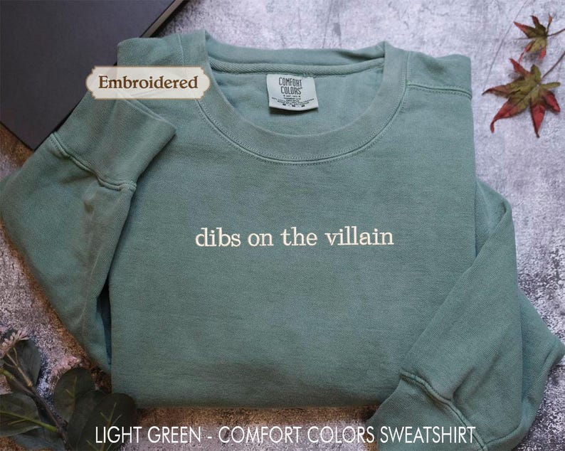 A light green, embroidered sweatshirt with the text "dibs on the villain" printed on it, placed on a gray surface with a leaf and a black object in the background.