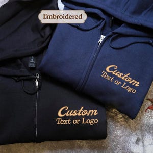 May include: Three zip-up hoodies in navy and black are shown. The navy hoodie has "Custom Text or Logo" embroidered in gold thread. The black hoodie also has the same text embroidered in gold. The word "Embroidered" is in a banner.
