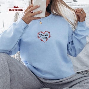 A person is sitting on a bed, wearing a light blue sweatshirt with a heart-shaped embroidered design and the text "Morally Grey" printed on it.