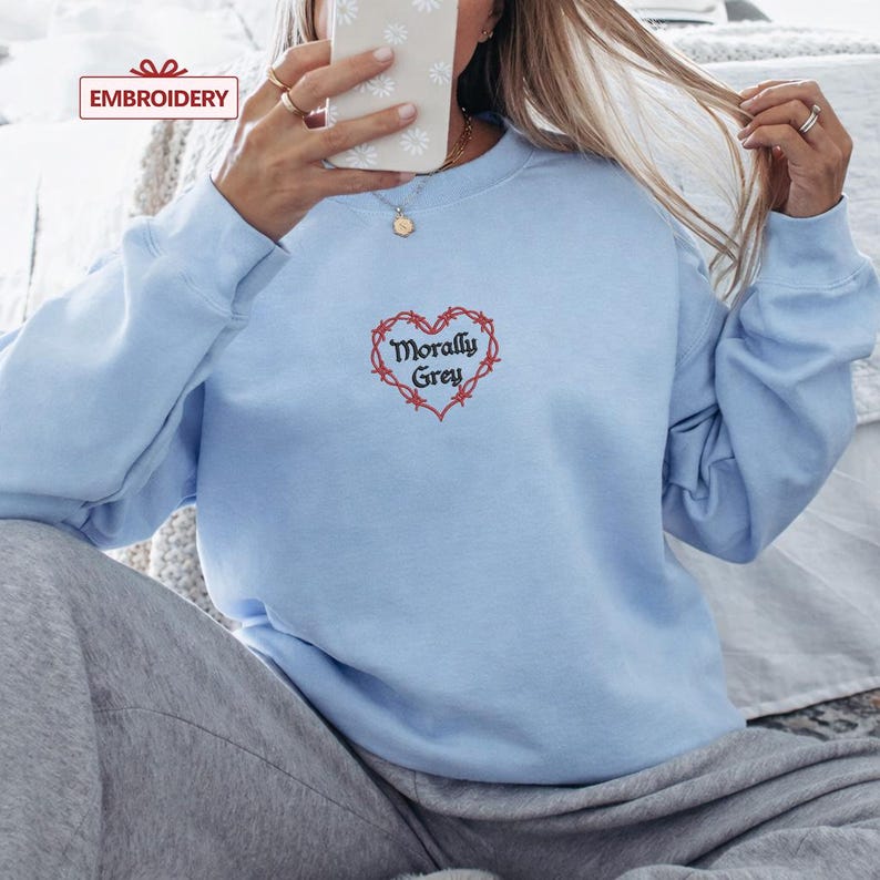 A person is sitting on a bed, wearing a light blue sweatshirt with a heart-shaped embroidered design and the text "Morally Grey" printed on it.