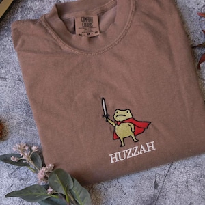 a brown t-shirt with a cartoon frog wearing a red cape and holding a sword, along with the word "HUZZAH" printed on it. The t-shirt is laid out on a gray surface, with a pine cone and some leaves nearby.