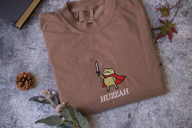 a brown t-shirt with a cartoon frog wearing a red cape and holding a sword, along with the word "HUZZAH" printed on it. The t-shirt is laid out on a gray surface, with a pine cone and some leaves nearby.