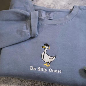 May include: A slate blue crewneck sweatshirt featuring an embroidered graphic of a white goose wearing a black graduation cap and a black necktie. Below the goose, the text Dr. Silly Goose is embroidered in white thread on the front of the garment.