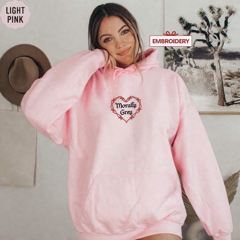 A woman wearing a pink hoodie with a heart-shaped embroidered design and the text "Morally grey" on it, posing in front of a wall with a framed picture.