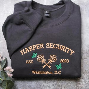 A black sweatshirt with gold text and graphics is displayed on a gray surface, accompanied by a small plant with white flowers.