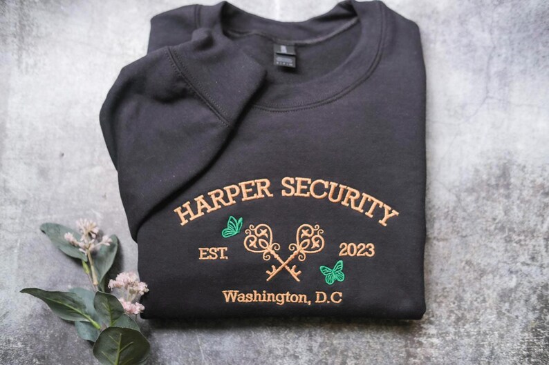 A black sweatshirt with gold text and graphics is displayed on a gray surface, accompanied by a small plant with white flowers.