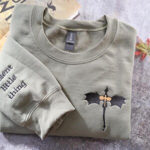 A gray sweatshirt with a dragon design and the text "Violent little thing" printed on the left sleeve, along with a book titled "The Darkest Thing" in the background.