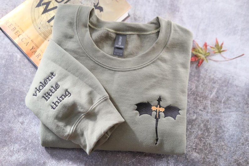 A gray sweatshirt with a dragon design and the text "Violent little thing" printed on the left sleeve, along with a book titled "The Darkest Thing" in the background.