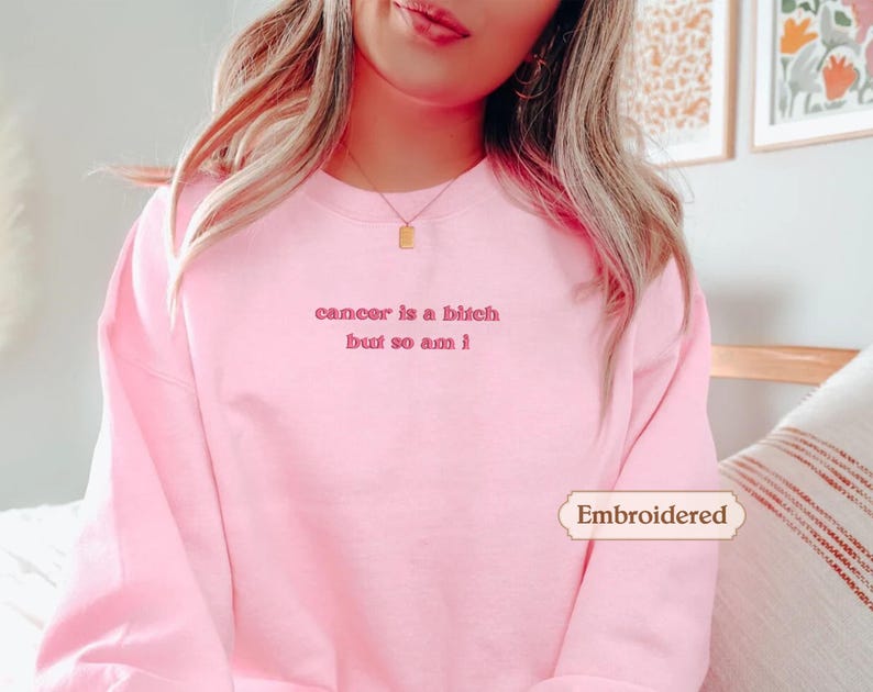 A woman wearing a pink sweatshirt with the text "cancer is a bitch but so am I" printed on it.