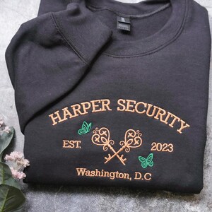 A black sweatshirt with embroidered text and graphics, including a key design, is displayed on a gray surface alongside a small plant with white flowers.