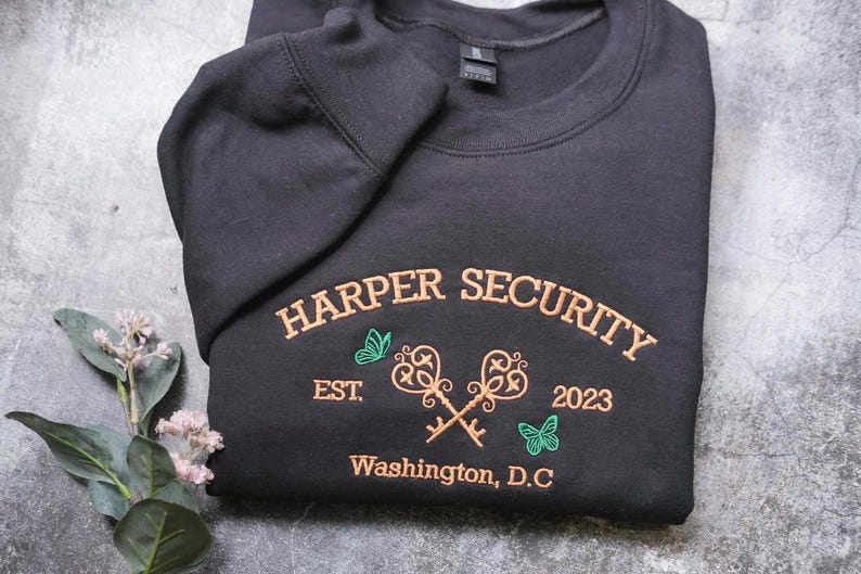 A black sweatshirt with embroidered text and graphics, including a key design, is displayed on a gray surface alongside a small plant with white flowers.