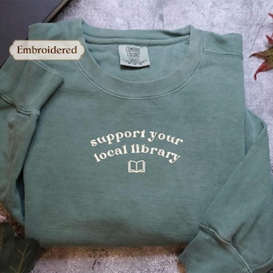 A light green long-sleeved shirt with the text "Support your local library" printed on it, along with a small book icon. The shirt is placed on a gray surface, with some leaves and a book visible in the background.