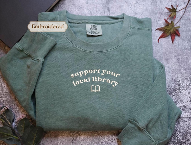 A light green long-sleeved shirt with the text "Support your local library" printed on it, along with a small book icon. The shirt is placed on a gray surface, with some leaves and a book visible in the background.