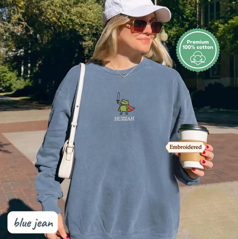A woman wearing a blue sweatshirt with a frog character and holding a coffee cup, standing on a brick sidewalk.