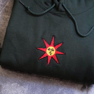 A black hoodie with a red sun embroidered on it, placed on a gray surface.