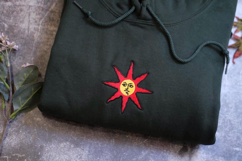 A black hoodie with a red sun embroidered on it, placed on a gray surface.