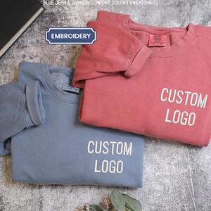 Peut inclure: Deux sweat-shirts à col rond de couleur bleu jean et cramoisi. Chaque vêtement présente le texte CUSTOM LOGO en lettres blanches sur le devant. Une petite étiquette rectangulaire bleue avec le mot EMBROIDERY est visible au-dessus du sweat.