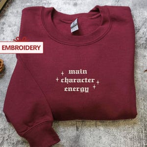 A burgundy sweatshirt with the text "main character energy" embroidered on it, along with a pine cone and autumn leaves on a gray surface.