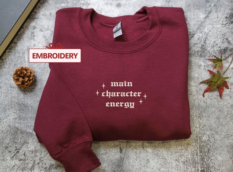 A burgundy sweatshirt with the text "main character energy" embroidered on it, along with a pine cone and autumn leaves on a gray surface.