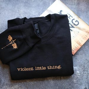 a black sweatshirt with a gold dragon design and the text "violent little thing" printed on it, along with a book titled "The King" lying next to it on a gray surface.