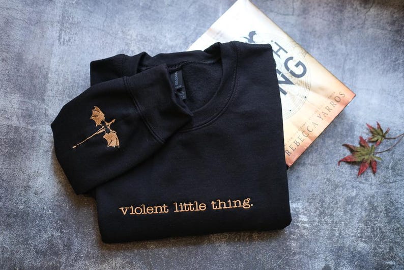 a black sweatshirt with a gold dragon design and the text "violent little thing" printed on it, along with a book titled "The King" lying next to it on a gray surface.