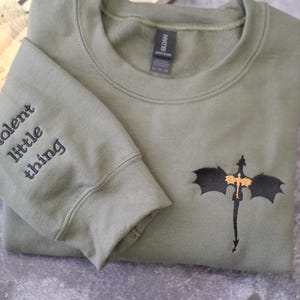 A grey sweatshirt with a dragon embroidered on the front, along with the text "Violent little thing" printed on the left sleeve.