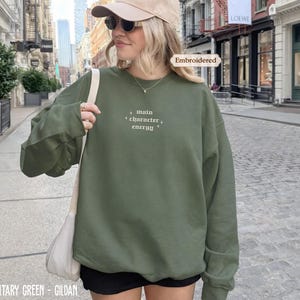 A woman wearing a green sweatshirt with embroidered text, sunglasses, and a cap is standing on a city street.